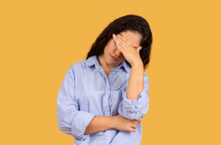 Photo for Stressed sad arab young woman in a light blue shirt holding her forehead in frustration or headache against a solid yellow background, with a pained expression on her face - Royalty Free Image