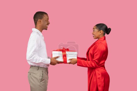 Photo for Smiling black man in shirt presents large white gift box with red ribbon to delighted woman in dress, capturing moment of joyful giving and surprise - Royalty Free Image