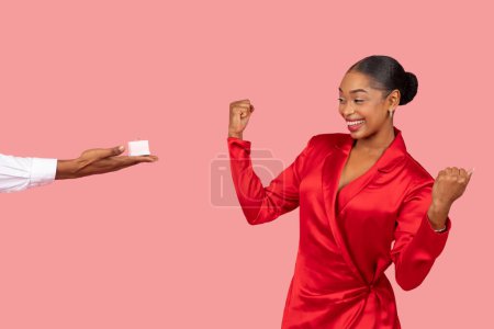 Téléchargez les photos : Femme noire ravie en robe rouge célèbre avec pompe poing que mans main présente une boîte de bague de fiançailles, créant un moment de surprise sur fond rose - en image libre de droit