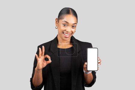 Téléchargez les photos : Glad jeune femme noire en costume formel montrer smartphone avec écran vide et ok signe avec la main, isolé sur fond de studio gris. Recommandation d'entreprise, application de travail, site Web - en image libre de droit