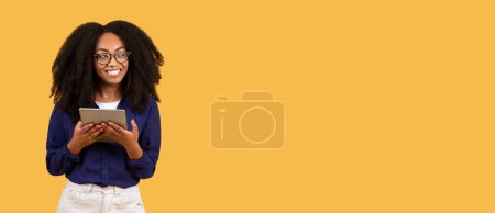 Foto de Entusiasta estudiante de negro en gafas, sosteniendo la tableta digital y mirando a un lado en el espacio de copia, colocada sobre fondo amarillo panorámico, pancarta - Imagen libre de derechos
