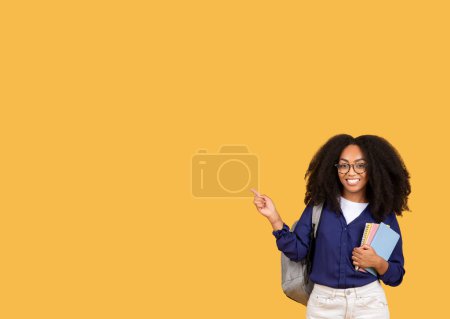 Photo for Cheerful black lady student, carrying backpack and copybooks, points to the side at copy space, set against lively yellow background, banner - Royalty Free Image