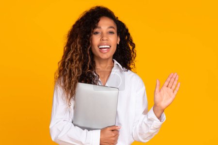 Téléchargez les photos : Joyeux afro-américaine dame tenant ordinateur portable et geste bonjour vous saluant, souriant à la caméra tout en posant sur fond jaune studio. Salut, j'ai une offre en ligne - en image libre de droit