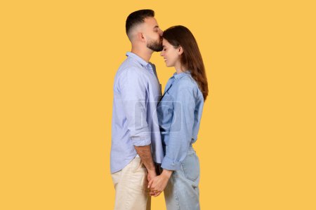 Photo for Romantic couple with eyes closed in tender forehead kiss, expressing affection and love, captured in a close embrace against a yellow background - Royalty Free Image