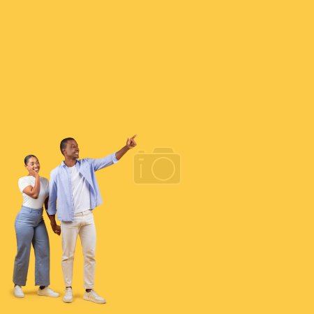Foto de Pareja negra entusiasta apuntando a la distancia en el espacio libre, con expresiones de alegría y anticipación, indicando algo interesante sobre fondo amarillo brillante - Imagen libre de derechos