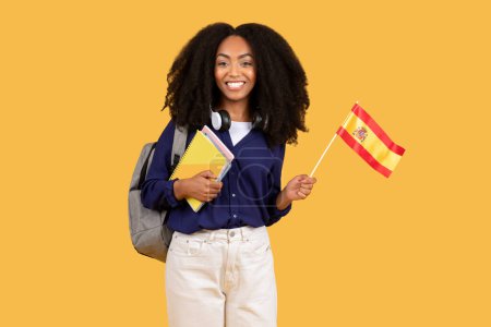 Photo for Happy black lady student, equipped with backpack and copybooks, proudly holds Spanish flag on yellow background, foreign language education and cultural exchange concept - Royalty Free Image