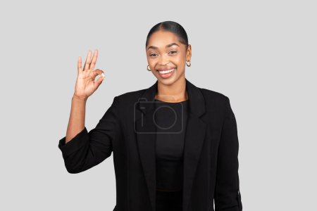 Téléchargez les photos : Radieuse femme d'affaires afro-américaine heureuse avec un sourire rayonnant donne un geste de main OK pour signifier l'approbation ou le succès, dans un contexte gris clair, studio. Style de vie professionnel - en image libre de droit