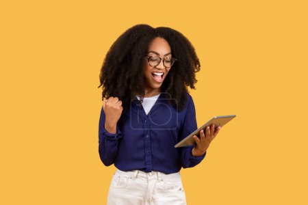 Elated young black woman with curly hair and glasses, holding tablet and making victory fist, exclaiming in triumph against yellow background
