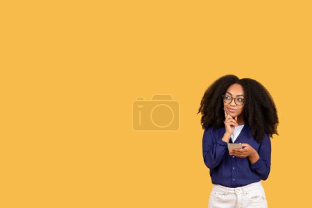Téléchargez les photos : Jeune femme noire réfléchie avec des cheveux bouclés et des lunettes tenant smartphone, regardant contemplatif, sur fond jaune uni, espace libre - en image libre de droit