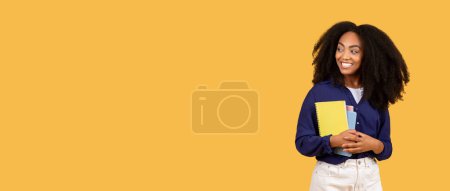 Téléchargez les photos : Joyeuse étudiante noire, tenant des copybooks et regardant de côté l'espace libre sur fond jaune, parfaitement préparée pour des publicités éducatives et des offres promotionnelles - en image libre de droit
