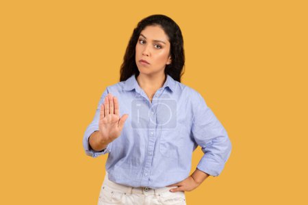 Serious confident arab young woman with curly hair, hand raised in a stop sign, looking at camera with a strong, assertive stance against a plain yellow background.
