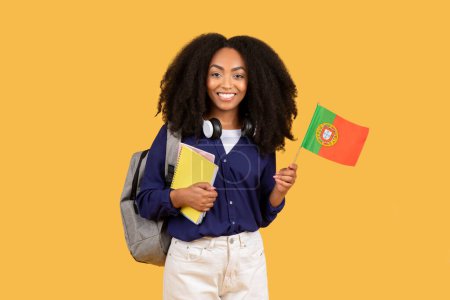 Foto de Mujer negra radiante con el pelo rizado, auriculares, portátiles y bandera portuguesa, exudando felicidad sobre fondo amarillo - Imagen libre de derechos