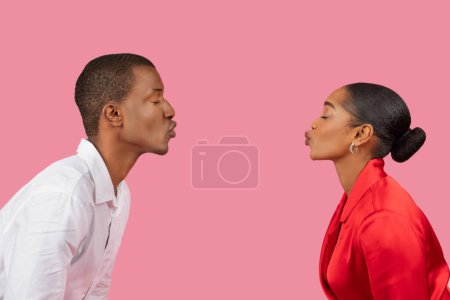 Foto de Vista del perfil del hombre y la mujer negros frente al otro con los labios fruncidos como si se besaran, creando una escena romántica sobre un fondo rosa sólido - Imagen libre de derechos