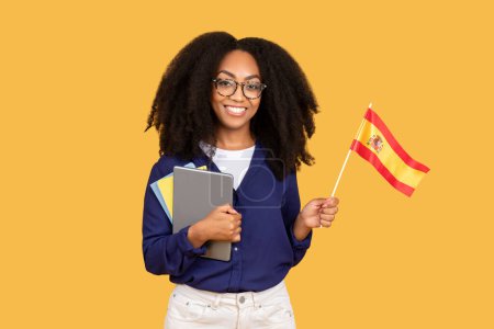 Téléchargez les photos : Joyeux étudiant noir, équipé de sac à dos et copybooks, tient fièrement drapeau espagnol sur fond jaune, symbolisant son enthousiasme pour la langue et la culture espagnole - en image libre de droit