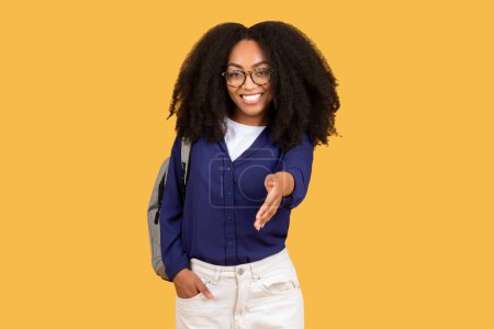 Téléchargez les photos : Accueillante jeune femme noire aux cheveux bouclés et lunettes étendant sa main pour une poignée de main, portant un sac à dos, sur fond jaune - en image libre de droit