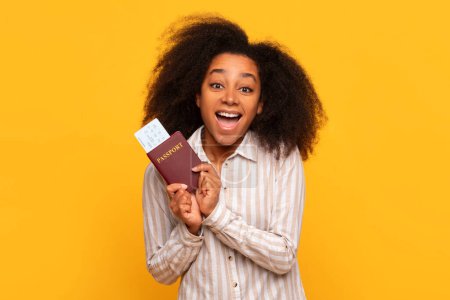 Téléchargez les photos : Joyeux jeune femme noire saisissant passeport avec carte d'embarquement, son visage allumé avec le frisson du voyage imminent, sur fond jaune ensoleillé - en image libre de droit