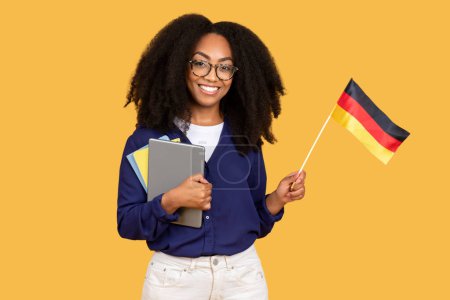 Téléchargez les photos : Étudiante noire joyeuse, tenant des copybooks et un drapeau allemand, rayonne de joie sur fond jaune, symbolisant son enthousiasme pour les études de langues étrangères et l'exploration culturelle - en image libre de droit