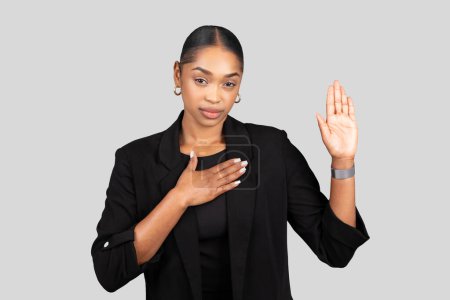 Foto de Mujer de negocios afroamericana sincera haciendo una promesa o jurando con la mano derecha levantada y la mano izquierda en el pecho, en un traje negro profesional sobre fondo gris, estudio - Imagen libre de derechos