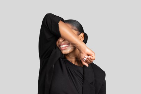Foto de Alegre mujer de negocios afroamericana alegre cubriéndose los ojos con un brazo, mostrando un lado espontáneo con una sonrisa radiante, vestida con atuendo profesional, estudio - Imagen libre de derechos
