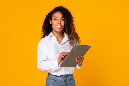 Téléchargez les photos : Joyeux Afro-Américaine s'engager avec tablette numérique pour le travail indépendant en studio sur fond jaune. Dame en chemise blanche décontractée heureusement websurfing tout en posant avec PC portable - en image libre de droit