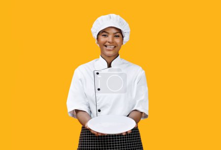 Foto de Chef femenino negro alegre orgullosamente presentando el plato blanco vacío en la cámara, cocinera afroamericana feliz invitando a una deliciosa comida, posando sobre un fondo amarillo soleado, espacio para copiar - Imagen libre de derechos