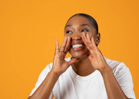 Téléchargez les photos : Joyeuse jeune femme afro-américaine tenant la main près de la bouche annonçant une excellente offre, regardant de côté sur fond de studio jaune. Bannière publicitaire avec espace libre - en image libre de droit