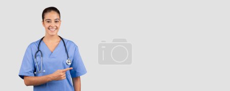 Foto de Amistoso profesional médico milenario caucásico en uniformes azules que apuntan a un lado, ideal para anuncios, con un estetoscopio, que incorpora experiencia y orientación en la atención médica - Imagen libre de derechos