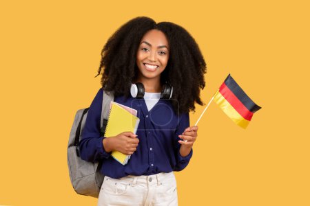 Foto de Brillante joven dama negra con el pelo rizado, auriculares, portátiles y bandera alemana, mostrando una sonrisa brillante sobre el fondo amarillo - Imagen libre de derechos
