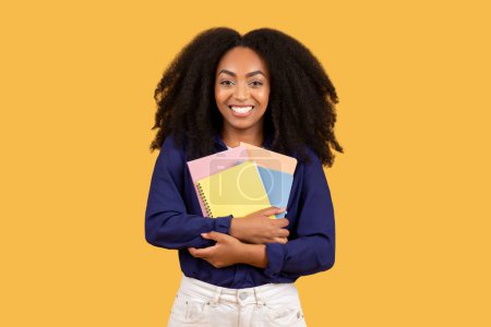 Photo for Cheerful black lady student, with beaming smile, holds copybooks looking at camera, set against vibrant yellow background, embodying positivity and academic enthusiasm - Royalty Free Image