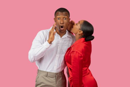 Photo for Black woman in red dress whispers secret into the ear of surprised man in white shirt, his hand over his mouth in shock, set against pink backdrop - Royalty Free Image