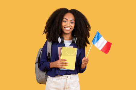 Foto de Estudiante afroamericana, con mochila y copybooks, sostiene bandera de Francia, posando sobre fondo amarillo, disfrutando de educación en idiomas extranjeros - Imagen libre de derechos