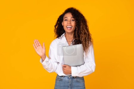 Téléchargez les photos : Jeune femme noire tenant son ordinateur portable fermé et saluant souriant à la caméra et vous saluant, debout sur fond de studio jaune. Services Internet publicité, offres en ligne - en image libre de droit
