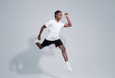 Téléchargez les photos : Homme noir dynamique en tenue de sport fonctionnant sur fond de studio gris, en mouvement plan de jeune homme afro-américain motivé présentant fitness et mode de vie actif, pleine longueur, espace de copie - en image libre de droit