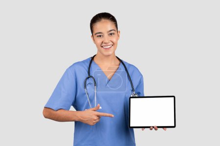 Téléchargez les photos : Professionnel de santé européen radieux dans des blouses bleues pointant vers une tablette avec un écran vierge, idéal pour les promotions d'applications médicales ou des informations de santé, isolé sur fond de studio gris - en image libre de droit