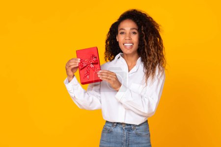 Téléchargez les photos : Joyeuse jeune femme afro-américaine tient une boîte-cadeau enveloppée posant sur fond jaune studio. Dame noire positive célébrant l'anniversaire ou les vacances, recevant un cadeau inattendu - en image libre de droit