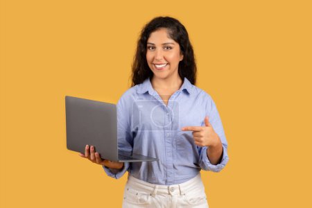 Foto de Mujer joven y amable con el pelo rizado sostiene una computadora portátil y señala a ella con una sonrisa, de pie con confianza sobre un fondo amarillo mostaza. Estudio y trabajo recomendación en línea - Imagen libre de derechos