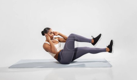 Fit black woman in sportswear doing abdominal bicycle exercise, making elbow to knee abs crunches on studio floor, over white backdrop, side view. Wellness and sporty lifestyle