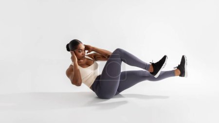 Photo for Perfect abs. Athletic young african american woman does elbow to knee crunches during fitness workout, flexing her abdominal muscles and core strength, lying over white studio backdrop. Panorama - Royalty Free Image