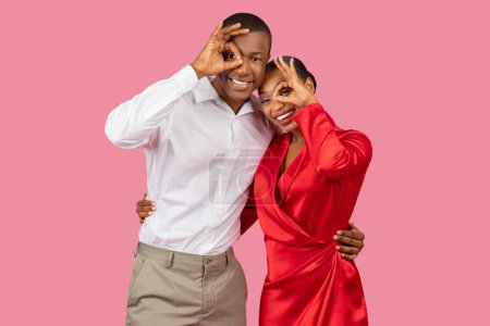 Photo for Playful black couple making circles around their eyes with their fingers, the woman in vibrant red dress and the man in crisp white shirt, both smiling on pink background - Royalty Free Image