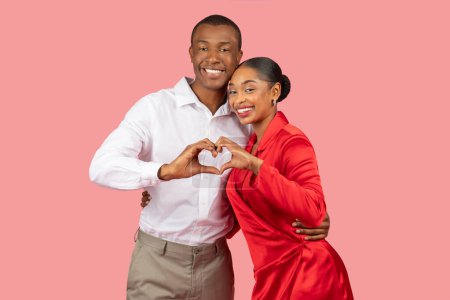 Smiling black couple in smart casual wear forming heart with their hands, symbolizing love and unity, set against soft pink background, radiating happiness
