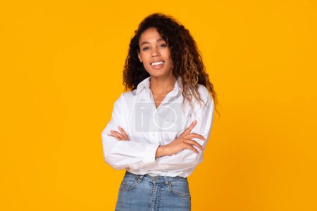 Photo for African American young woman in white shirt and casual denim, poses crossing hands with cheerful smile and confidence, standing against yellow studio backdrop, looking at camera - Royalty Free Image