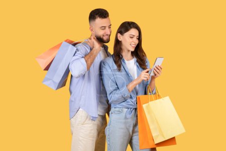Photo for Happy spouses holding shopper bags and using smartphone, couple browsing mobile application with sale offers and discounts, standing over yellow background - Royalty Free Image