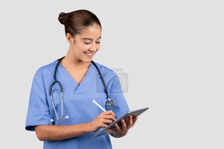 Téléchargez les photos : Professionnel médical du millénaire caucasien engagé dans des blouses bleues à l'aide d'une tablette numérique, accédant éventuellement aux renseignements sur le patient ou aux dossiers médicaux, avec un stéthoscope autour du cou - en image libre de droit