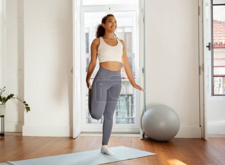 Young fitness lady in sportswear working out in the morning, making heel to butt exercise stretching leg in living room interior, starting workout training with warmup. Healthy sport. Full length