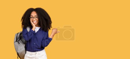 Téléchargez les photos : Étudiante souriante jeune femme noire aux cheveux bouclés et aux lunettes pointant vers un espace latéral vide, parfaite pour la publicité, avec sac à dos sur fond jaune, panorama - en image libre de droit