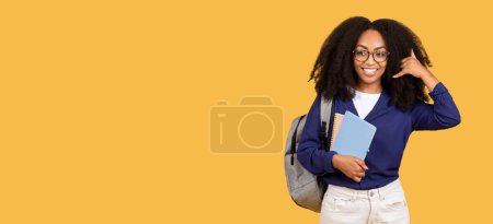 Photo for Cheerful black lady student, equipped with copybooks and backpack, playfully gestures call me, set against yellow background with free space, panorama - Royalty Free Image