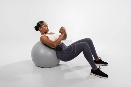 Téléchargez les photos : Femme américaine africaine sportive faisant des exercices abdominaux sur le ballon de remise en forme, s'entraînant sur fond de studio blanc. Fit sportive noire travaillant sur ses muscles de base et la force du corps - en image libre de droit