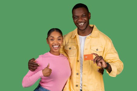 Photo for Happy Black Couple Holding Credit Card, Showing Thumb Up, Recommending Service Smiling To Camera Over Green Background. Excited Bank Customers Showing Card For Payments Posing In Studio - Royalty Free Image