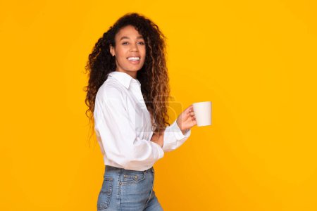 Foto de Feliz joven afroamericana sosteniendo taza de café y sonriendo a la cámara en el fondo amarillo del estudio. Preciosa dama negra en camisa blanca haciendo descanso con bebida caliente. Espacio libre - Imagen libre de derechos