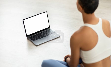 Téléchargez les photos : Femme latine engagée assise sur le sol à la maison en tenue d'entraînement, utilisant un ordinateur portable éventuellement pour un cours de fitness ou une séance d'entraînement personnelle, mélangeant technologie et exercice - en image libre de droit